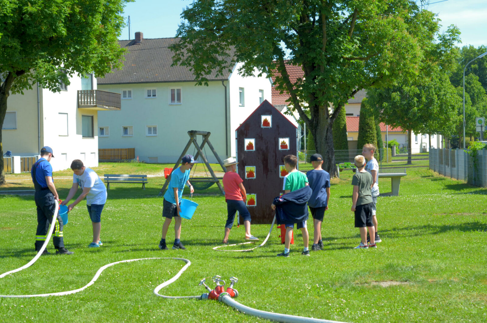 Kinder löschen spielerisch ein simuliert brennendes kleines Haus auf einer Wiese, unterstützt von einem Feuerwehrmann.