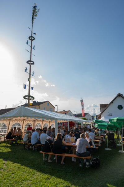 Viele Personen sitzen auf Bierbänken unter einem Zelt. Im Hintergrund der Maibaum.